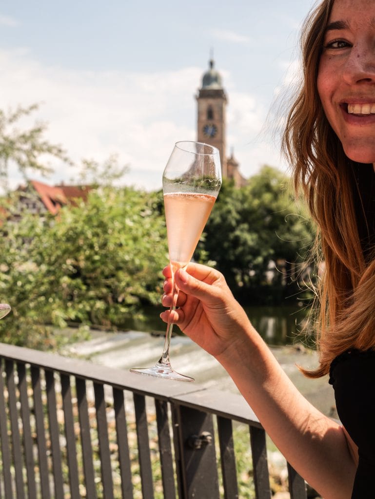 Frau hält ein Glas Rosé-Sekt am Neckar in Nürtingen, im Hintergrund die Stadtkirche St. Laurentius, Symbol für Genuss und Lebensfreude im Belsers Hotel