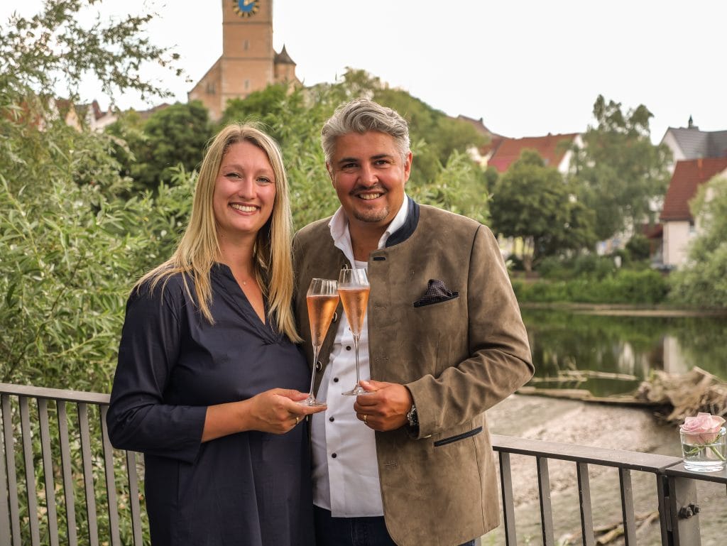 Zwei Personen stoßen mit Sektgläsern an, im Hintergrund die Stadtkirche St. Laurentius in Nürtingen, Symbol für Gastfreundschaft und Hotelgründung