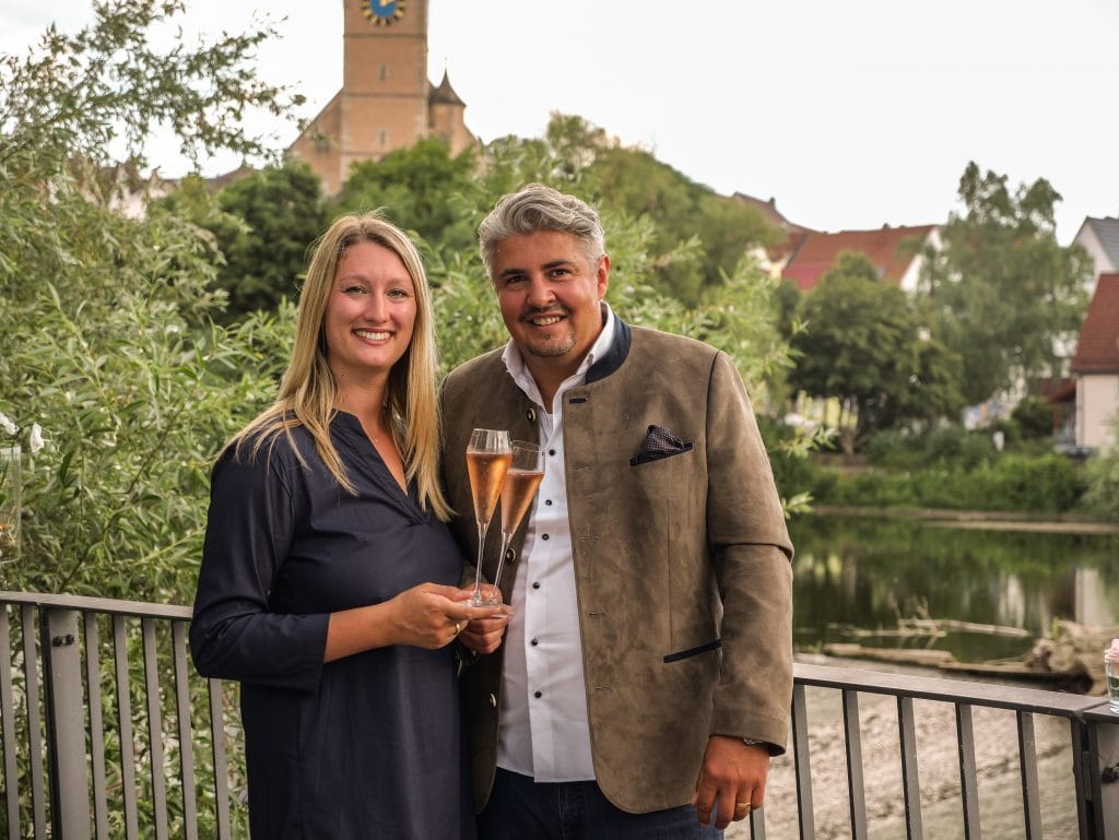 Zwei Personen stoßen mit Sektgläsern an, im Hintergrund die Stadtkirche St. Laurentius in Nürtingen, Symbol für Gastfreundschaft und Hotelgründung