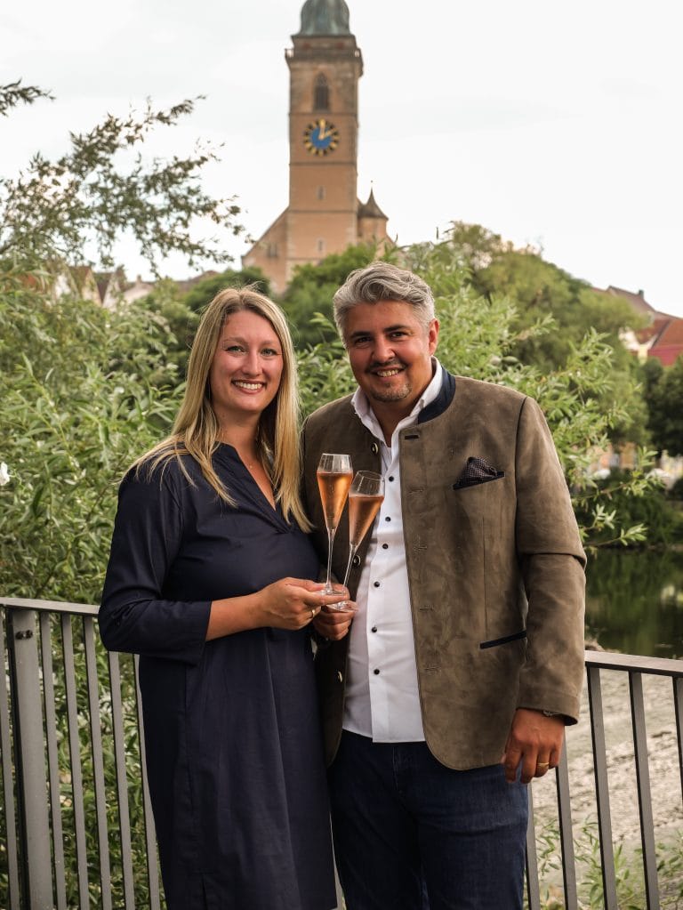 Zwei Personen stoßen mit Sektgläsern an, im Hintergrund die Stadtkirche St. Laurentius in Nürtingen, Symbol für Gastfreundschaft und Hotelgründung