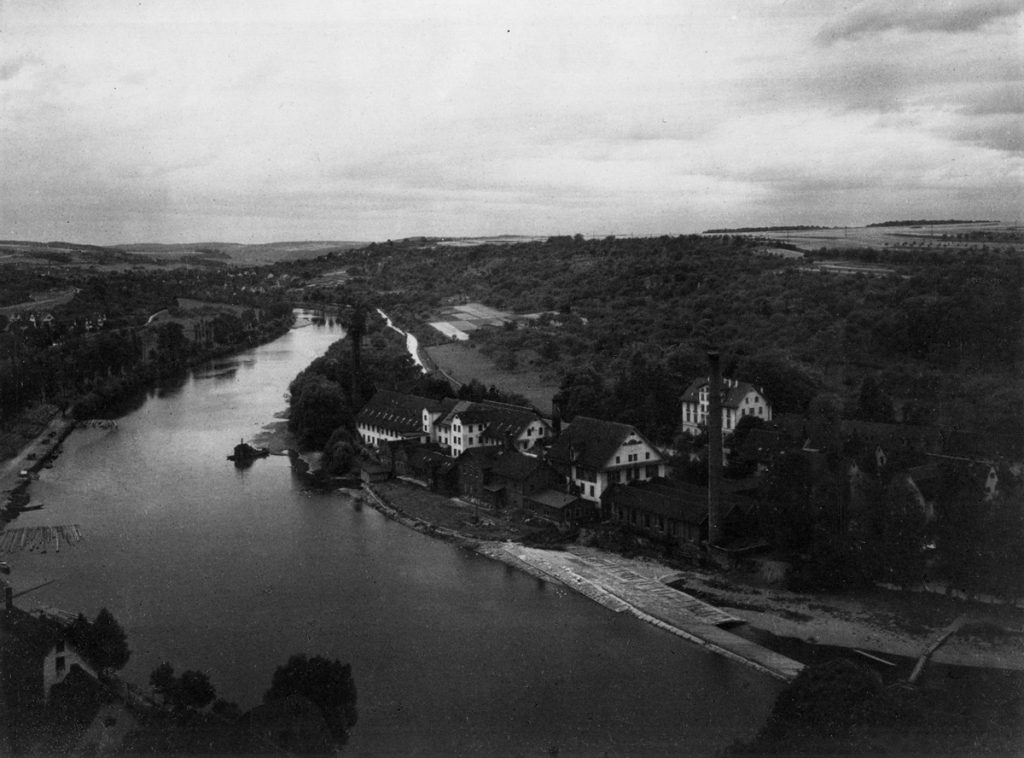 Historisches Schwarz-Weiß-Foto von Belsers Hotel am Flussufer in einer ländlichen Umgebung mit Fluss, Bäumen und alten Gebäuden.