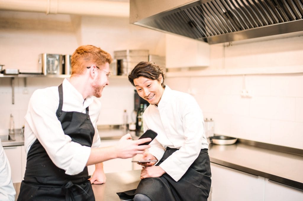 Ein junges Kochteam in einer modernen Hotelküche, das gemeinsam am Handy lacht und Spaß hat, symbolisiert Teamgeist und kulinarische Exzellenz im Belsers Hotel.
