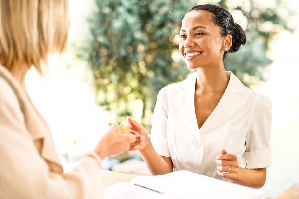 Begrüßungsszene im Belsers Hotel bei Sonnenschein, freundliche Rezeptionistin beim Empfang, hochwertiger Service, Entspannung, komfortable Unterkunft, perfekter Aufenthalt, Schlüsselübergabe.