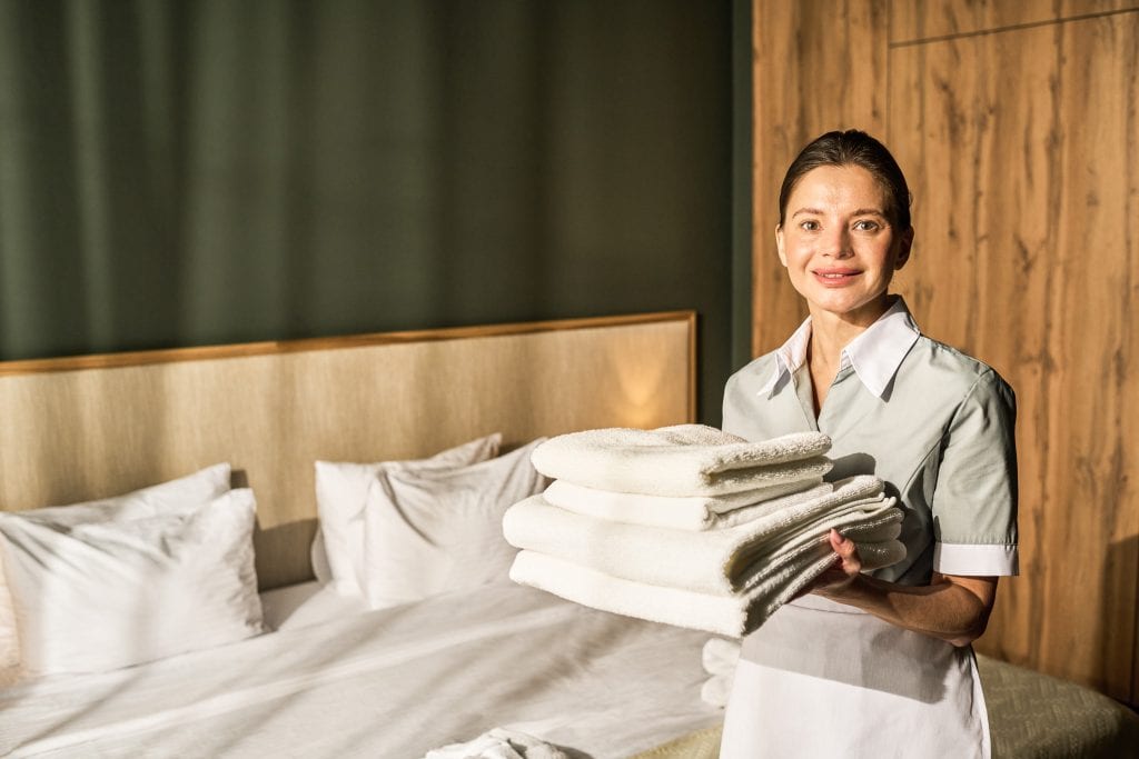 Erschlossene Bilder - eine Hotelangestellte hält frisch gewaschene Handtücher in einem modernen Hotelzimmer, zeigt freundlichen Service, Komfort und Sauberkeit, ideal für Urlaubs- oder Geschäftsreisende.