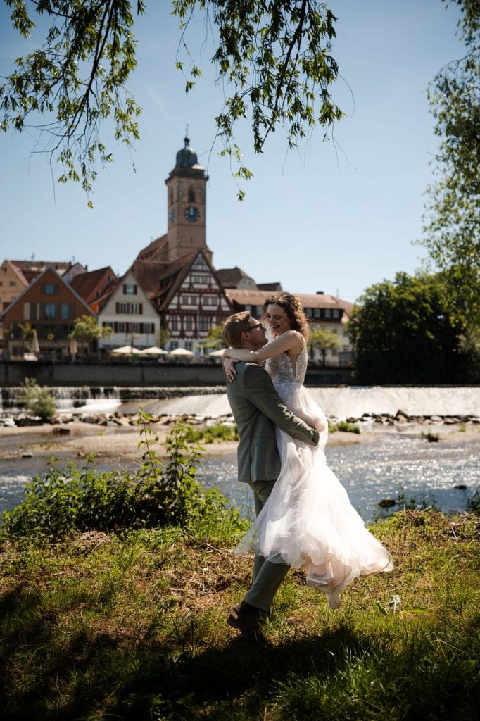 Braut und Bräutigam tanzen am Flussufer in Nürtingen bei Sonnenlicht, mit historischem Stadtkern im Hintergrund.